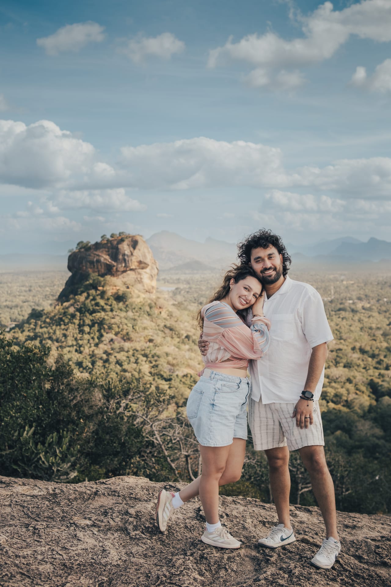 A couple holding each other on a hilltop with Sigiriya rock and the plains behind them