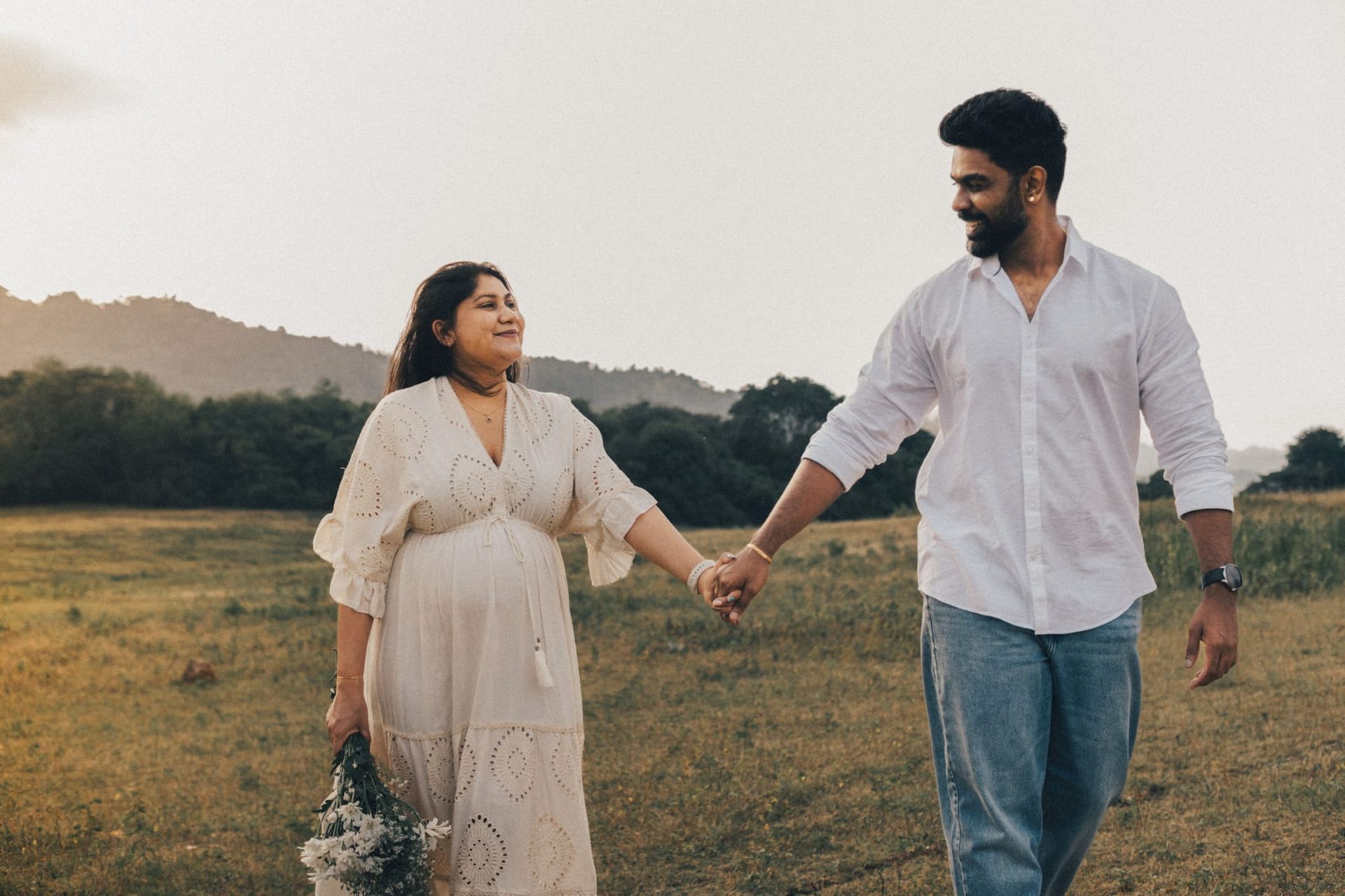 A couple walking hand in hand through a dry field at dusk, she in a flowing cream dress, the hills behind them