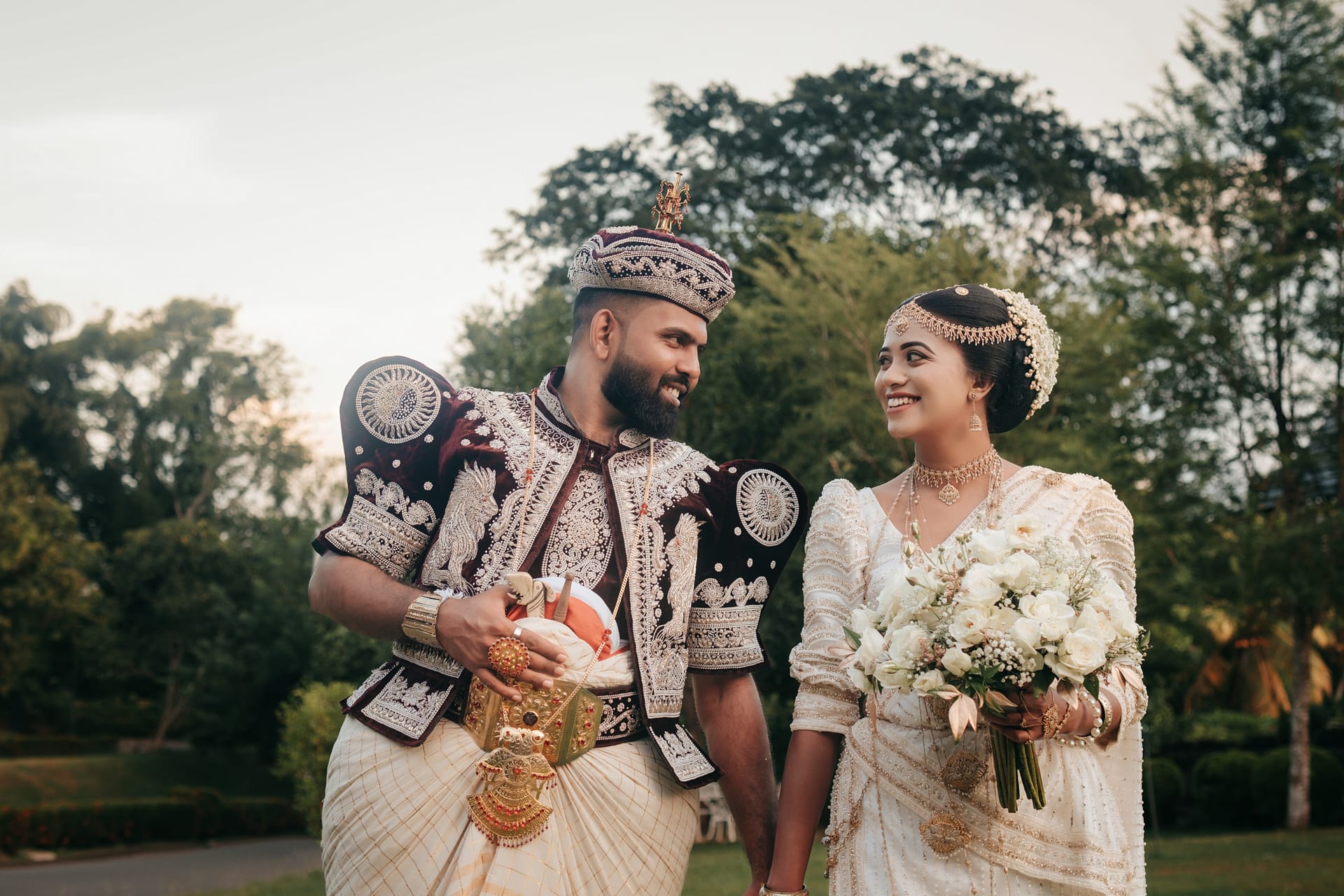 A bride and groom in full Kandyan dress walking together through a garden