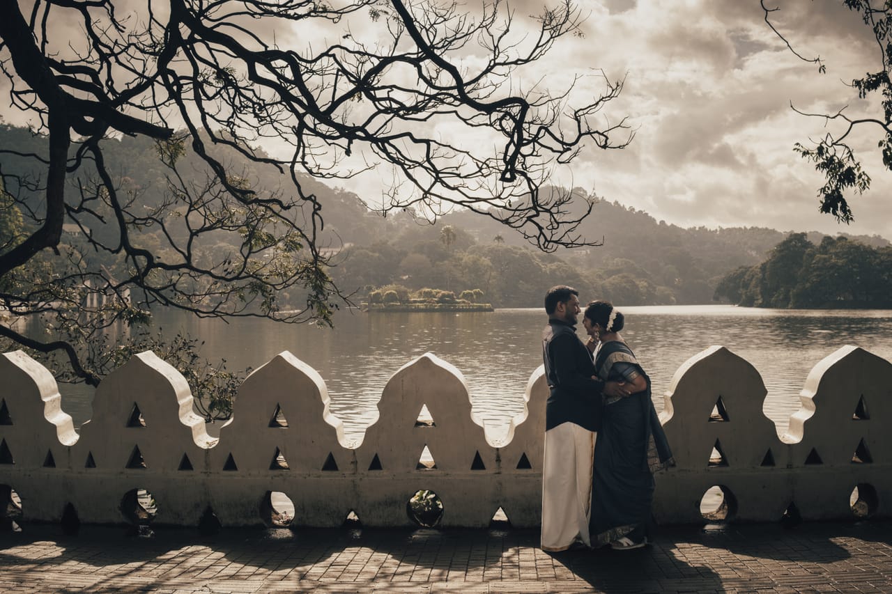 A couple standing at a lakeside pavilion in Kandy, framed by bare branches
