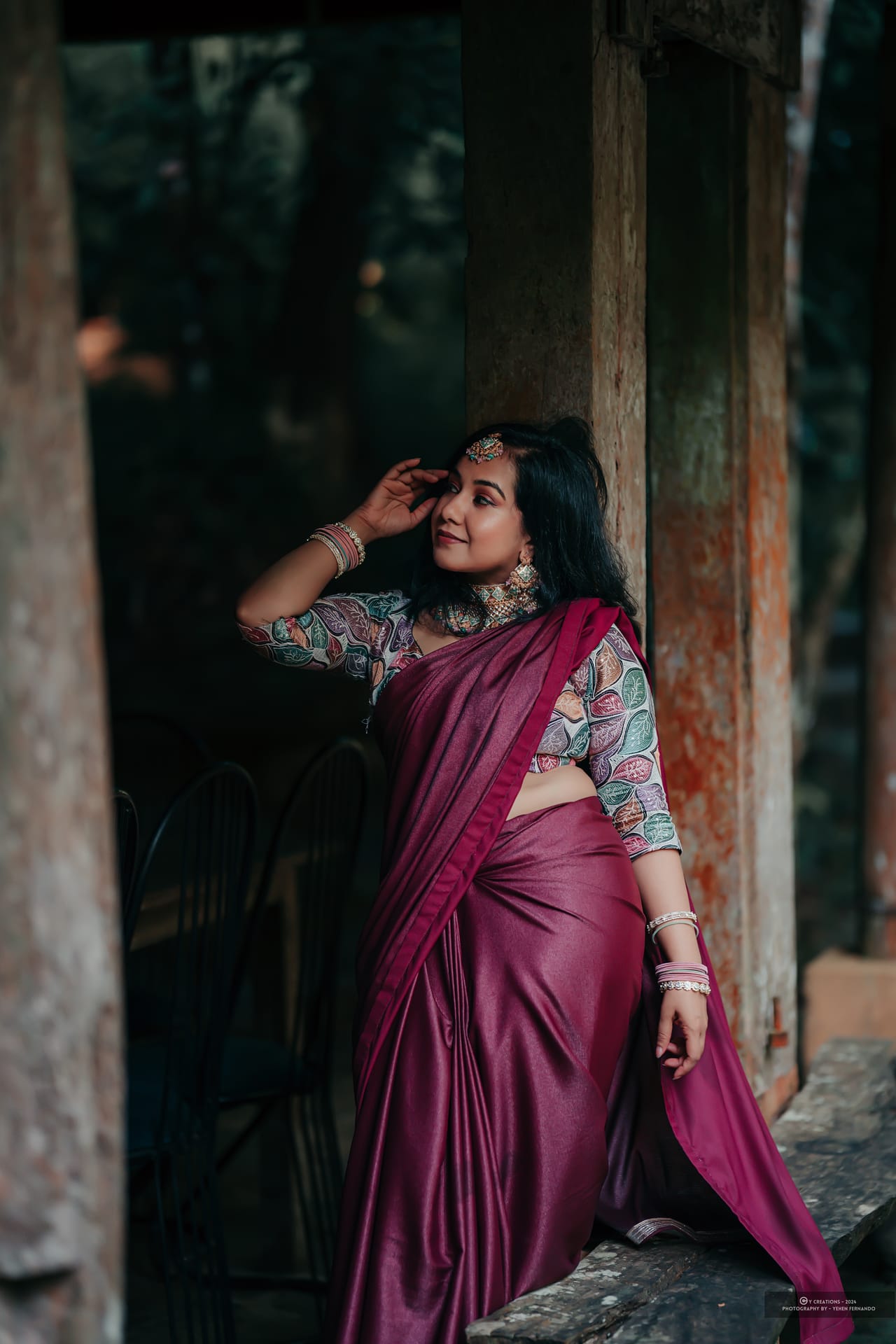 A woman in a maroon saree lit softly against a dark colonial interior