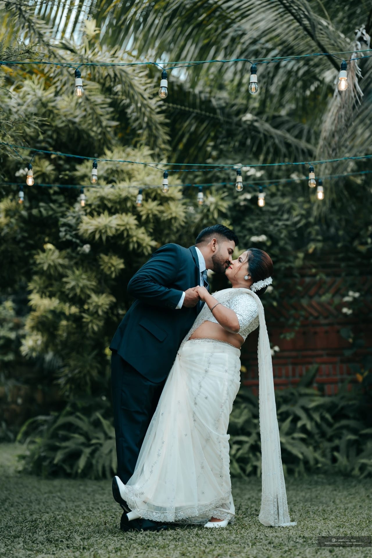 A groom dipping his bride for a kiss under strings of warm bulbs in a garden