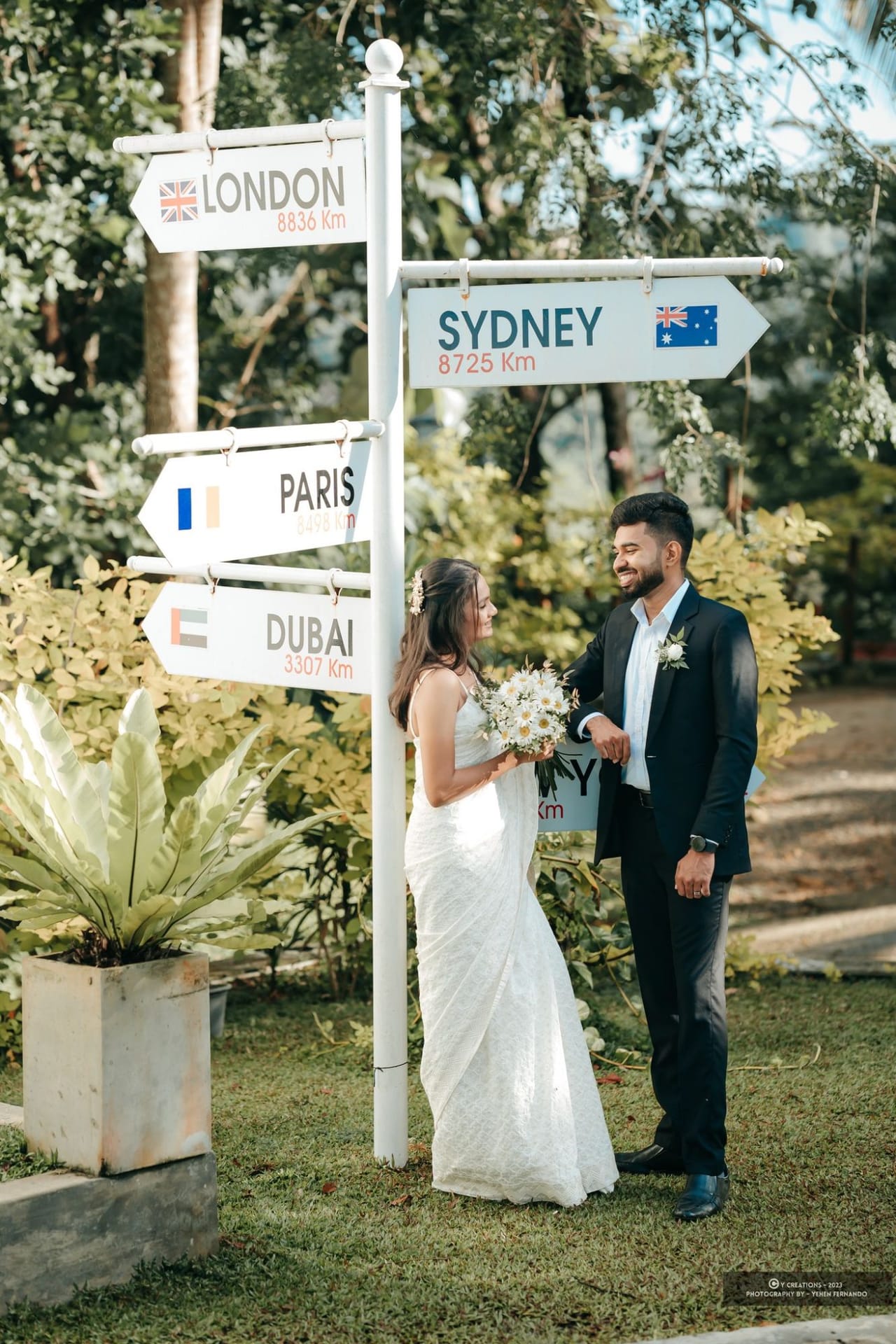 A couple laughing beneath a garden signpost pointing to London, Sydney, Paris and Dubai