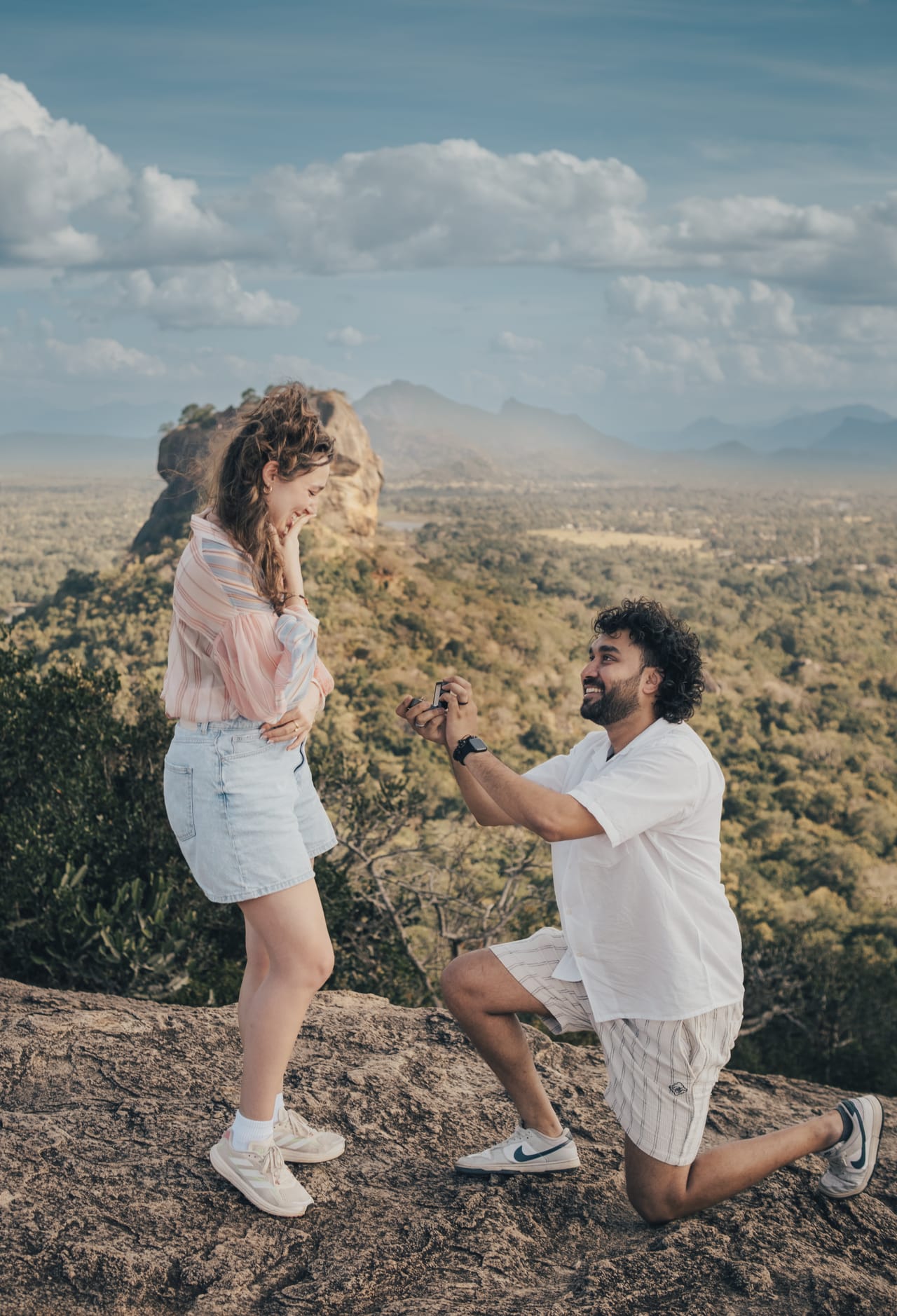 A man on one knee proposing at sunrise, his partner's hands to her face, Pidurangala behind them