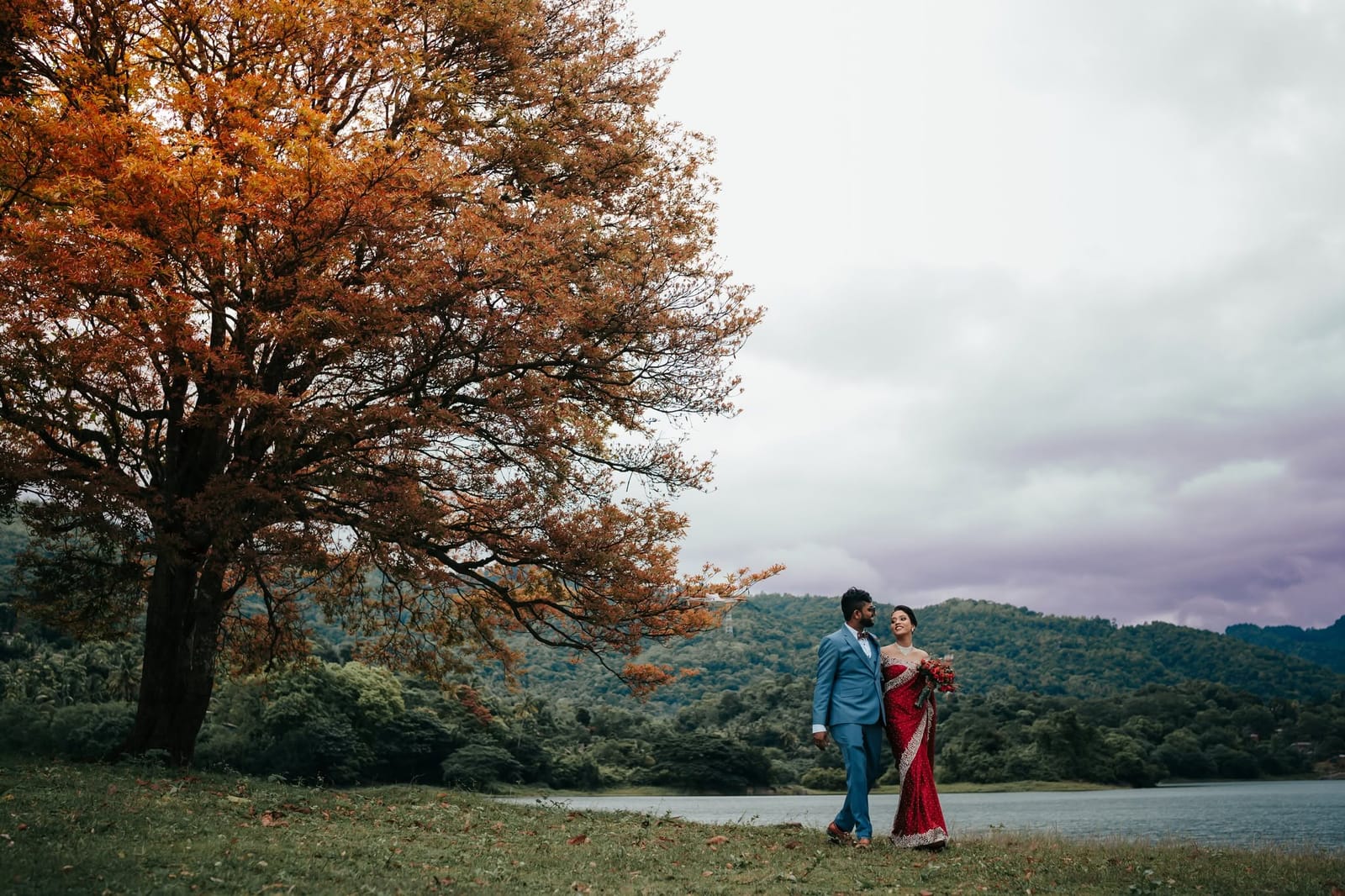 A couple walking by a lake beneath a tree turning colour