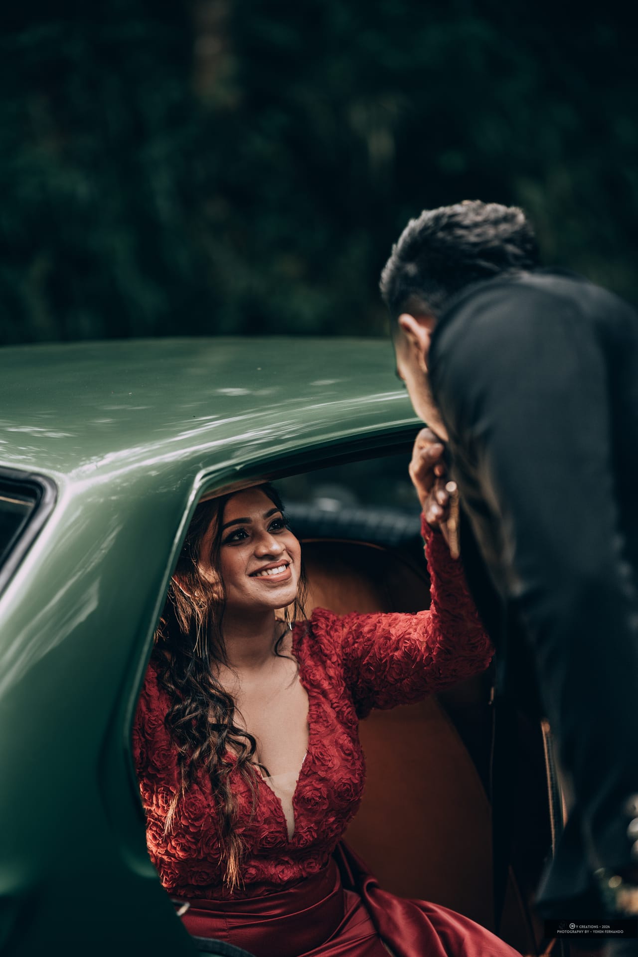 A woman in a red dress leaning from a vintage green car during a pre-shoot