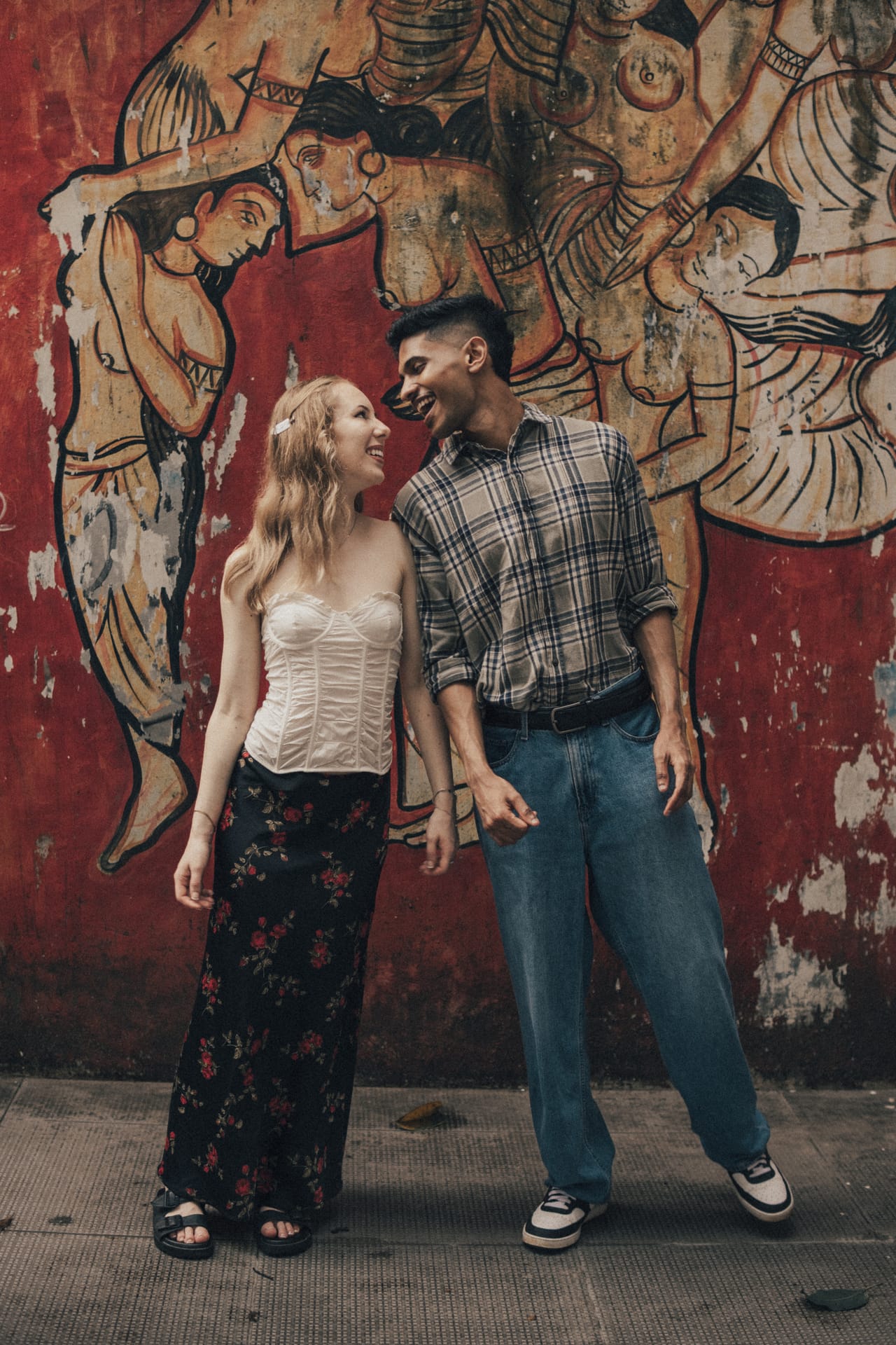 A travelling couple laughing together in front of a painted red mural wall