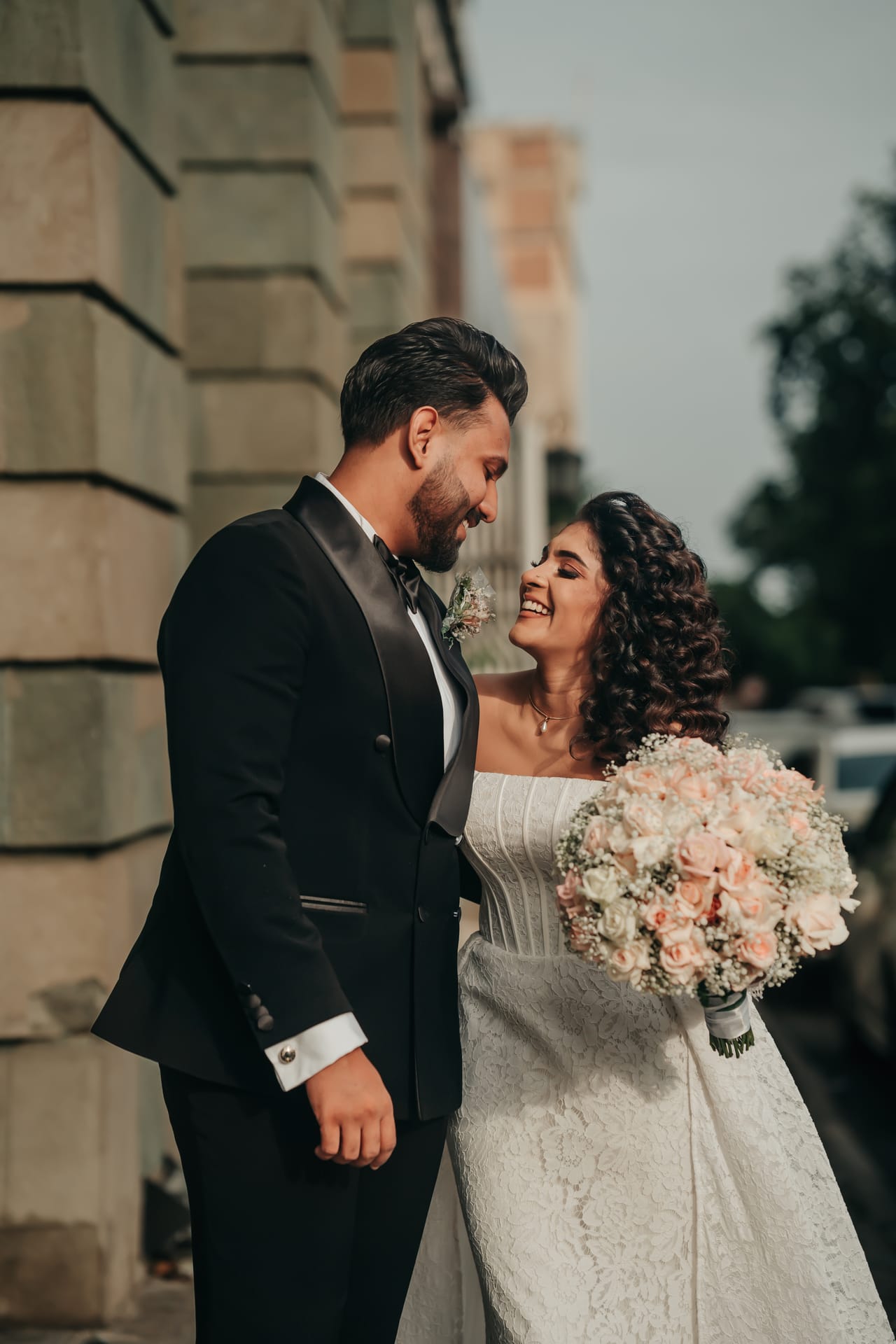 A groom in a black tuxedo laughing with his bride in a white gown holding her bouquet